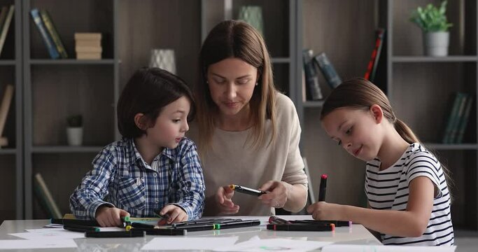 Caring young mother or nanny teaching little kids drawing pictures in paper album. Happy small children siblings involved in creative activity together with mum on weekend at home, daycare concept.