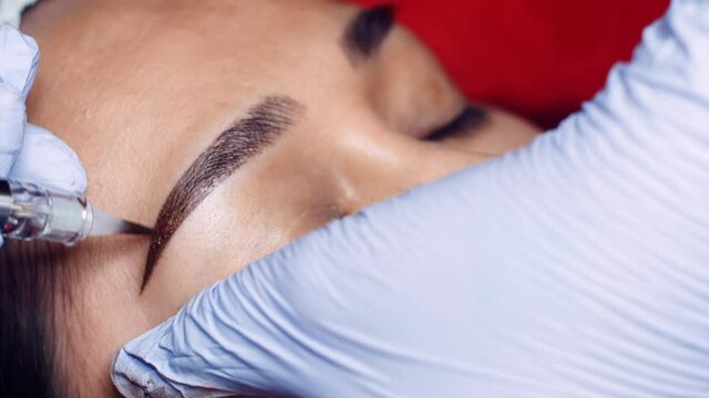 Close up microblading eyebrow tattoo. Beautician in gloves makes permanent makeup correction of the shape of the eyebrows to a young women. Eyebrow tattoo in a beauty salon.