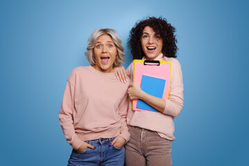 Two caucasian women are prepared for studies holding the necessary books. Portrait of astonished girls. Posing on a blue background