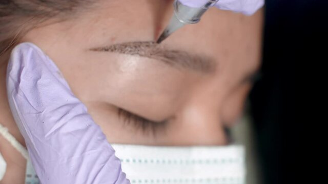 Close up microblading eyebrow tattoo. Beautician in gloves makes permanent makeup correction of the shape of the eyebrows to a young women. Eyebrow tattoo in a beauty salon.