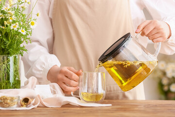 Woman pouring tasty chamomile tea from teapot into cup on table
