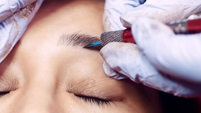 Close up microblading eyebrow tattoo. Beautician in gloves makes permanent makeup correction of the shape of the eyebrows to a young women. Eyebrow tattoo in a beauty salon.