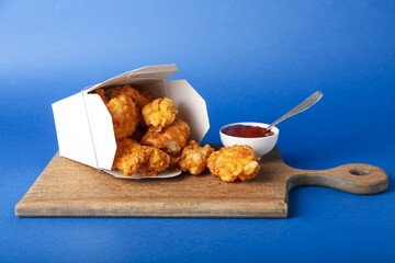 Board with tasty fried popcorn chicken and tomato sauce on blue background