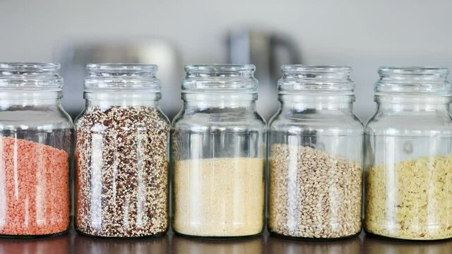 clear pantry jars with different types of grains and legumes including quinoa  lentils buckwheat and barley on benchtop with kitchen bokeh, simple ingredients