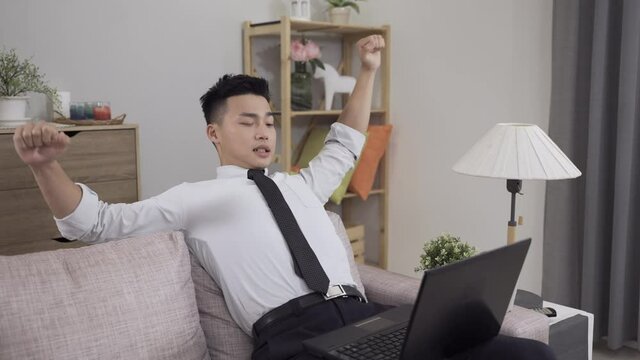 asian guy working from home is continuing typing on computer after leaning back with an arm stretch and exercising his stiff neck on the sofa in the living room