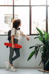 Young multiracial college student with backpack and red skateboard looks out the window. Copy space. Vertical image. © Daniel
