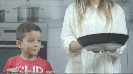 Beautiful young blonde girl with her kid, tossing pancake in the kitchen.