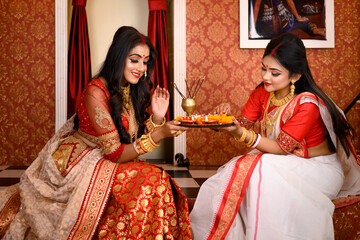 Two Indian women wearing traditional saree, gold jewellery and bangles sitting at home with flower and light diyas in decorative background. Indian festival, culture, occasion, religion and fashion. - obrazy, fototapety, plakaty
