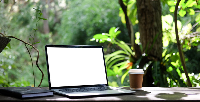 Laptop computer, coffee cup and notebook on wooden table with nature background.