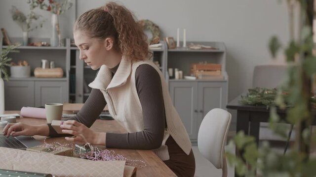Medium long of young curly-haired Caucasian woman sitting at desk in her craft shop, looking at small gift boxes and using laptop computer, preparing for sending packages to customers