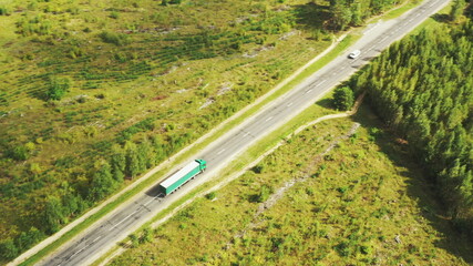 Aerial View Of Highway Road Through Field And Summer Green Forest Landscape. Top View Of Truck Tractor Unit Prime Mover Traction Unit In Motion On Freeway. Business Transportation, Trucking Industry.