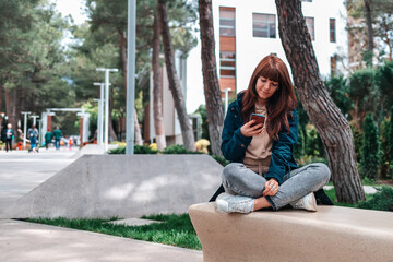 Fototapeta premium A young beautiful woman sits cross-legged on a park bench and uses a smartphone. Outdoor. Free wi-fi area