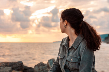 A young woman sits alone by the sea and looks at the sunset. View from the back. The concept of...