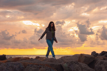A young woman walks alone on the sea rocks. The dramatic sunset in the background. The concept of...