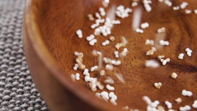 Popped dry amaranth falls into a rustic wooden bowl in slow motion on a jute cloth. Puffed pseudocereal grains. Macro