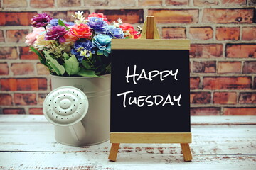 Happy Tuesday text on black wooden easel and vase with flowers bouquet on brick wall background