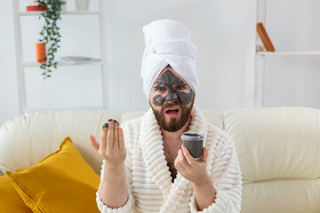 Bearded funny man having fun with a cosmetic mask on his face made from black clay. Men skin care,...