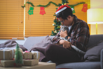 Young man wearing Santa hat sitting on couch with his cat enjoying spending time together in Christmas time at home.