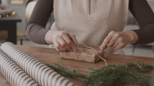 Close-up of cropped craftsperson sitting at desk in workplace, decorating small present box with cut pieces of Christmas tree branches and tying them with twine