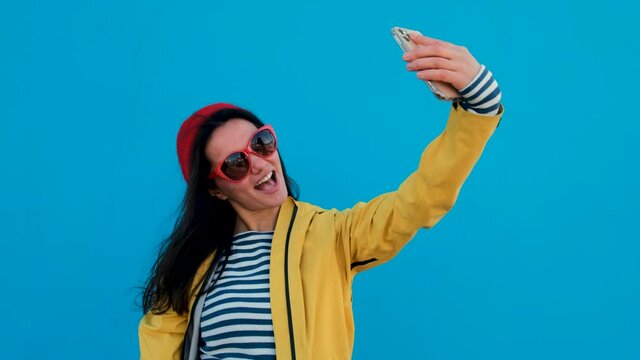 Portrait of young woman taking a selfie by smartphone wearing a knitted white hat on colorful blue background