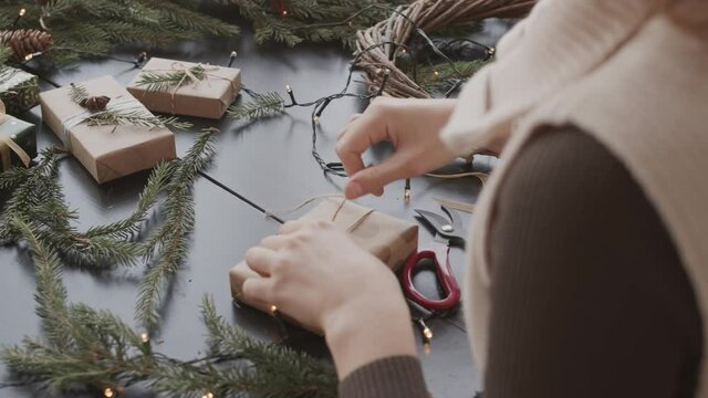 High angle over-shoulder of cropped craftsperson sitting at desk in workplace, decorating small present box and tying them with twine