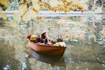 young beautiful plus size model in autumn brown warm coat and hat sitting in boat on pond decorate with chrysanthemums and apples in autumn park, concept of season leaf fall, romance and unusual date