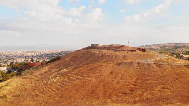 Aerial rising view Erebuni fortress ruins on hilltop with Yerevan city panorama background . Old urartian fortified city . Important historical site in Armenia