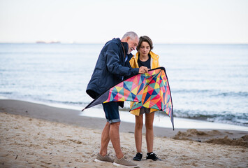 Senior man and his preteen granddaughter preparing kite for flying on sandy beach.