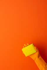 A hand in a protective rubber glove holds a yellow washing sponge on a orange background