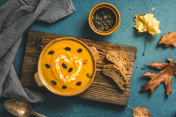 Bowl with pumpkin soup on wooden board on blue background, top view. Decorated with punpmkin seeds and autumn leaves.