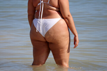Overweight woman in white swimsuit standing in a sea water. Vacation on a beach, overeating and...