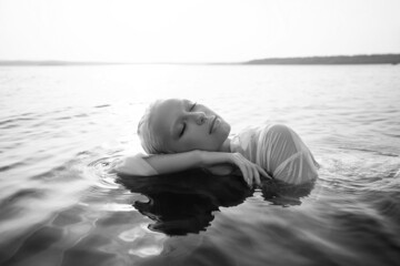Fototapeta premium Beautiful blonde woman short hair lies in water on sea shore, pond. Art portrait of a relaxing woman in the water on shore
