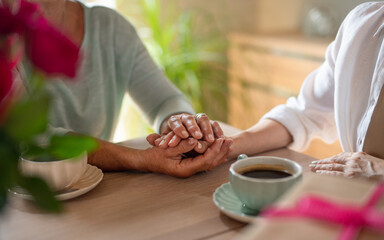 Close up of senior mother holding hand of adult daughter when having coffee together indoors at home