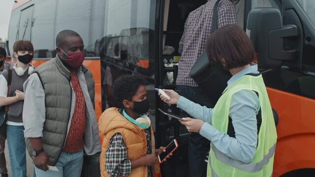 Medium shot of female bus supervisor in acid green vest and face measuring temperature of passengers with contactless infrared thermometer while they getting on bus