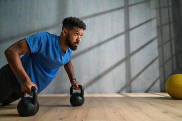 Young African American sportsman doing push ups with kettle bells indoors, workout training concept.