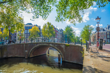 Fototapeta premium Beautiful sunny day on the old street of Amsterdam. Narrow canal, old bridge and traditional houses. Sunlight. Amsterdam, Holland, Netherlands, Europe.