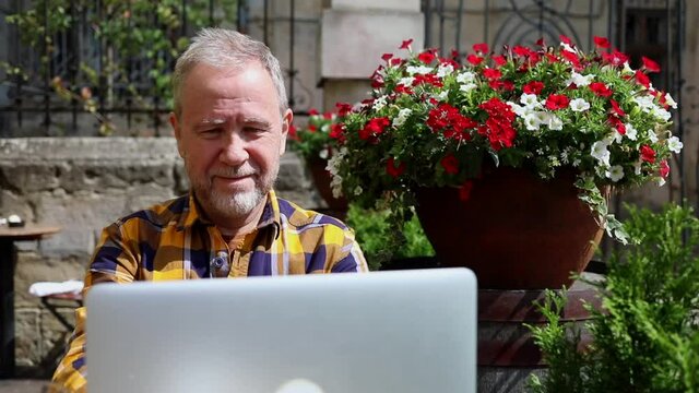 Smiling adult bearded man using modern laptop outside enjoying sunny beautiful day resting in cafe outside. Internet connecting. Communicating online chat app. Traveling free urban lifestyle.