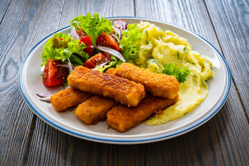 Fried fish sticks with potato puree and fresh vegetable salad on wooden table
