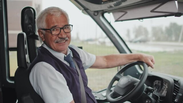 Medium portrait of cheerful senior bus driver in uniform sitting at driver seat with hands on steering wheel smiling to camera