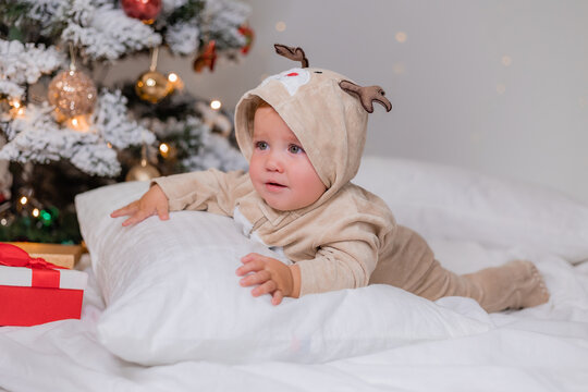 cute chubby cheeked baby in a jumpsuit with deer horns lies on a large white pillow under Christmas tree. winter New Year's concept. space for text. High quality photo