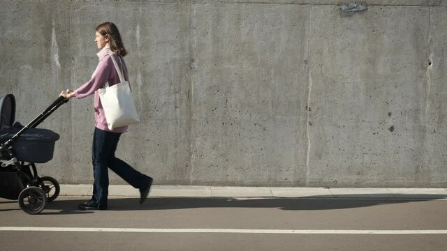 Mother with stroller and canvas eco shopping bag walking near grey concrete wall in the city. Caring mom looking on newborn in baby carriage. Zero waste parenthood.