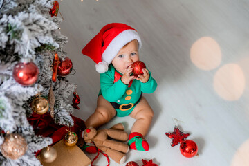 cute baby in a green bodysuit and a Santa hat decorates the Christmas tree with colorful balloons. child in a Christmas gnome costume. winter New Year's concept. space for text. High quality photo
