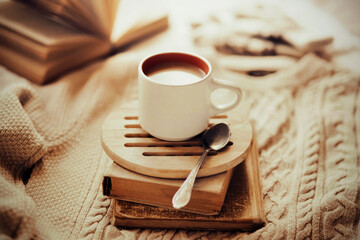 On a stack of old books there is a cup of cocoa with milk and a teaspoon. The still life is located on a beige wool patterned sweater on a bright morning. Home comfort in the cold season. Hygge.