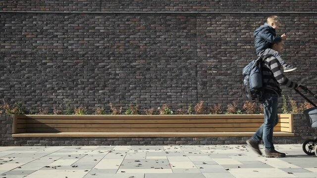 Father carrying his little son on shoulders and pushing newborn baby stroller near brick wall. Happy man in maternity leave with pram and child.