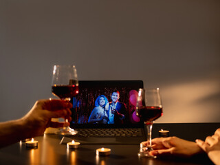 Home party. Romantic evening. Virtual friends greeting. Two glass of wine in hands toasting on laptop screen with friendly congrats in festive candles decorated room interior.