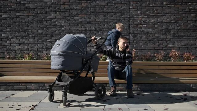 Busy father with newborn baby stroller sitting near brick wall and talking by smartphone. Man in maternity leave with pram and child trying to work.