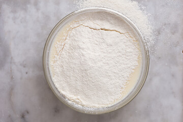 Top view of glass bowl with batter in it covered by sifted flour on marble surface