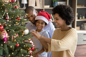 Happy mixed race little kid boy decorating Christmas tree with smiling bonding young african...