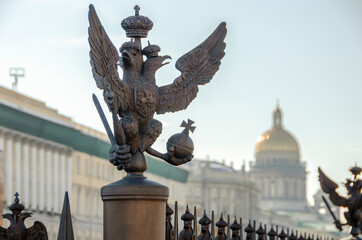 Fototapeta na wymiar Double-headed eagle on the fence of the Alexander Column at the Winter Palace and a view of St. Isaac's Cathedral (blurred focus), St. Petersburg, Russia