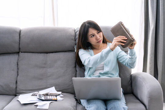 Concerned stressed young Asian woman sitting on sofa at home looking her empty purse with paper letters from bank informing about debt bankruptcy. Financial, unprofitable, debt concept.